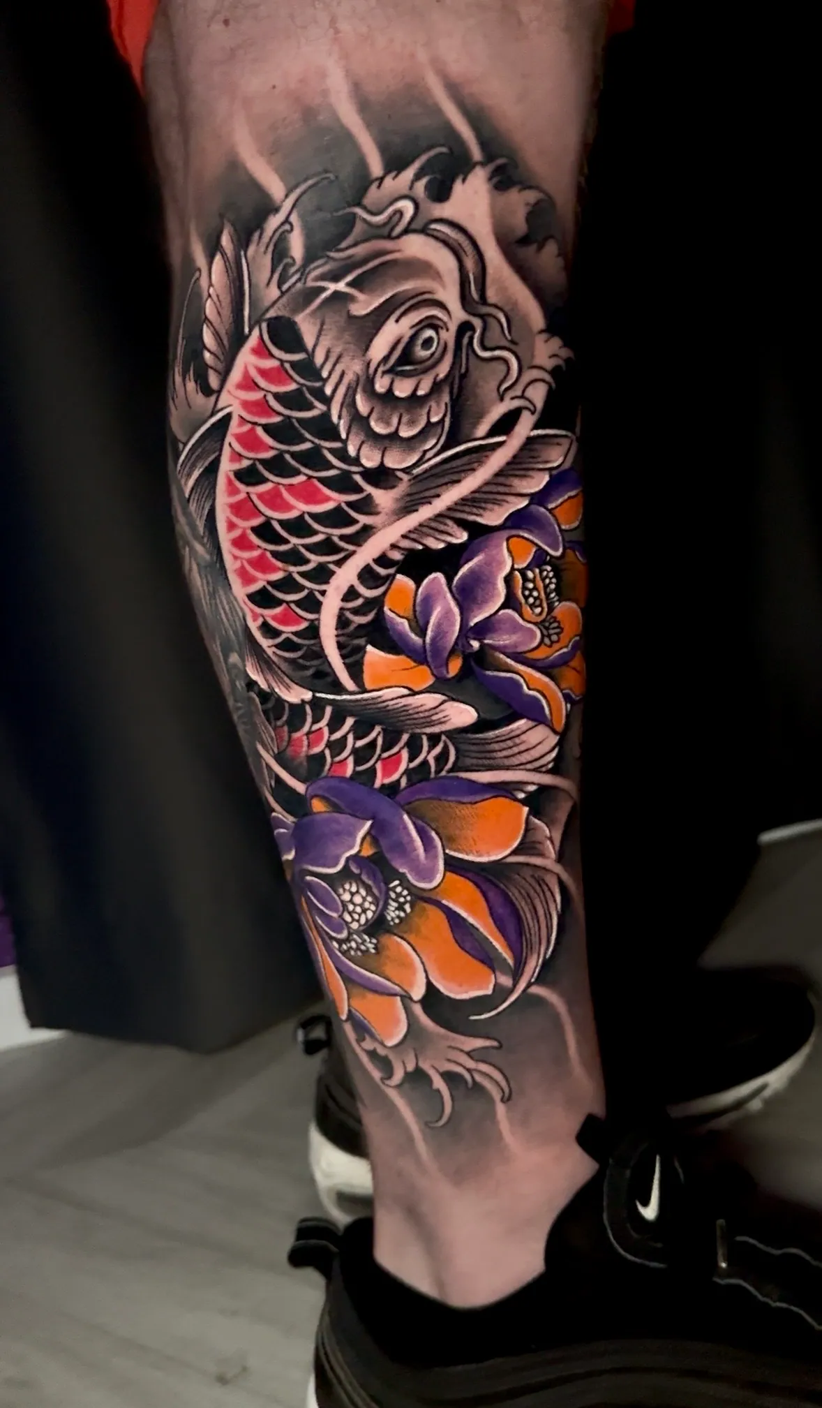Tetovaža u boji koi ribe sa ljubičastim i narandžastim cvetovima na potkolenici, Dropz Tattoo Studio Niš